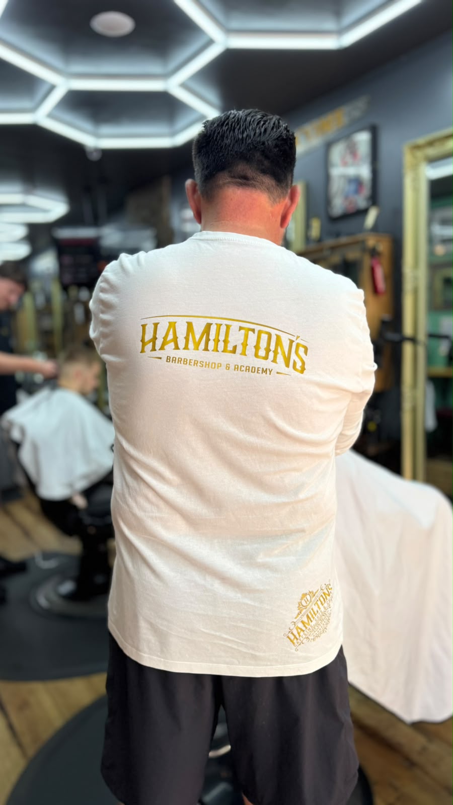 A Hamilton's barber in a gold-branded white t-shirt on the shop floor while a cut is underway behind him